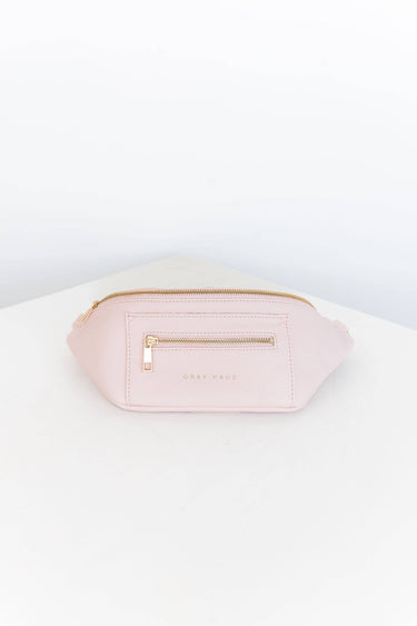 A light pink belt bag with gold zippers is centered on a white surface. The bag features two zippered compartments on the front, and the words